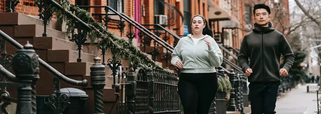 Dos personas corriendo en una acera de una ciudad junto a casas de ladrillo.